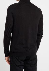 Svart polo-tröja med en formad design, ribbad textur vid kragen och ärmsluten, detaljerade raglanärmar, bärs över mörka byxor.