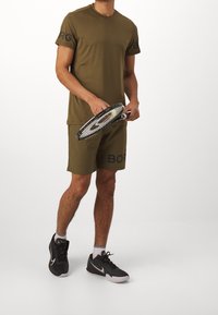 Olivově zelené sportovní tričko s černými akcenty, doplněné o sladěné šortky s nápisem "BORG." Černé tenisky s bílými podrážkami a pruhované ponožky.