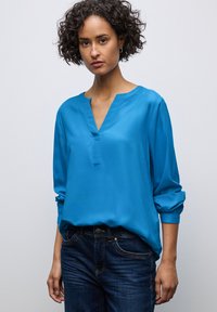 Turquoise blouse met een V-hals en lange mouwen, gemaakt van gladde stof. Gecombineerd met blauwe denim jeans met zakken en stikseldetails.