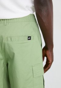 Svetlo zeleni cargo hlače imajo teksturirano tkanino, odprto zadnjo žep in črno oznako Nike na hrbtni strani.