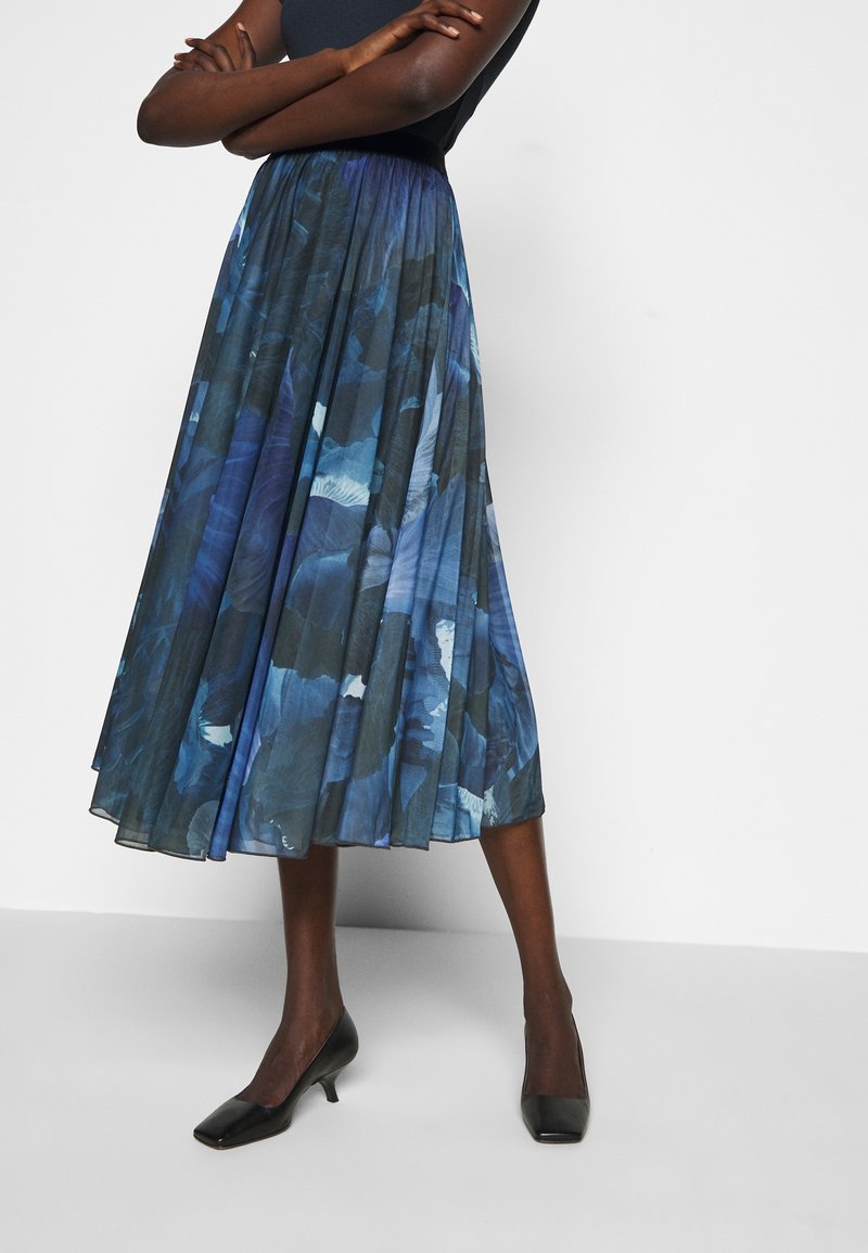 Kvinna klädd i en blå och svart blommönstrad plisserad midikjol i kombination med svarta låga klackskor med fyrkantig tå, stående med armarna i kors.