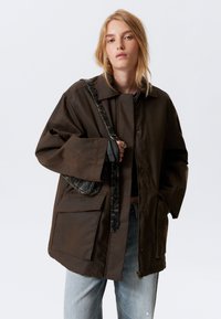 Brun lynlåsjakke med store forlommer og vide ærmer. Fremstillet sammen med en tekstureret sort og grå taske med et mønstret skulderbånd.