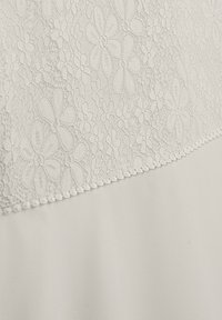 Spitzenpaneel mit floralem Muster in sanftem Beige oben, ergänzt durch einen glatten, einfarbigen beigen Stoff darunter und dekorative gezackte Abschlüsse.