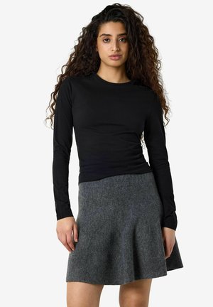 Jeune femme aux longs cheveux bouclés portant un top noir à manches longues et une jupe grise en ligne A, debout devant un fond clair uni.