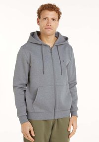 Felpa grigia con cerniera, realizzata in tessuto morbido, dotata di cappuccio con coulisse, tasche frontali e un piccolo logo sul petto.