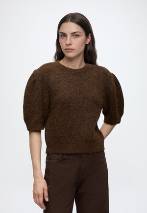 Femme aux longs cheveux bruns porte un pull marron duveteux à manches courtes bouffantes et un pantalon marron foncé sur un fond blanc uni.
