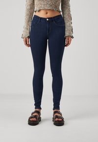Dunkelblaue Slim-Fit-Jeans mit glattem Denim-Textur, Fünf-Taschen-Design und Knopfverschluss vorne, kombiniert mit klobigen schwarzen Sandalen.