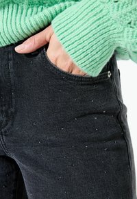 Jeans en denim noir avec un détail de poche et de petits embellissements brillants, associés à un pull en tricot côtelé vert clair.