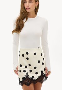 Femme portant un haut à manches longues blanc et une mini jupe blanche à pois noirs avec un bord en dentelle noire, debout devant un fond uni.