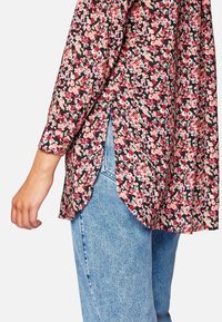 Blusa floreale con fondo nero, fiori rosa e crema, maniche lunghe, spacco laterale e dettaglio a bottoni sui polsini.