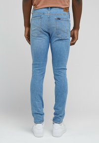 Jeans in denim azzurro chiaro con vestibilità slim, caratterizzati da tasche posteriori, cuciture a contrasto e un'etichetta marrone in vita. Indossati con sneakers bianche.
