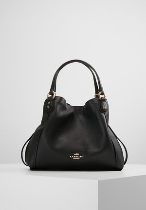 Bolso Coach de cuero negro con doble asa, herrajes dorados y emblema con logo, exhibido sobre una superficie blanca contra un fondo gris claro liso.
