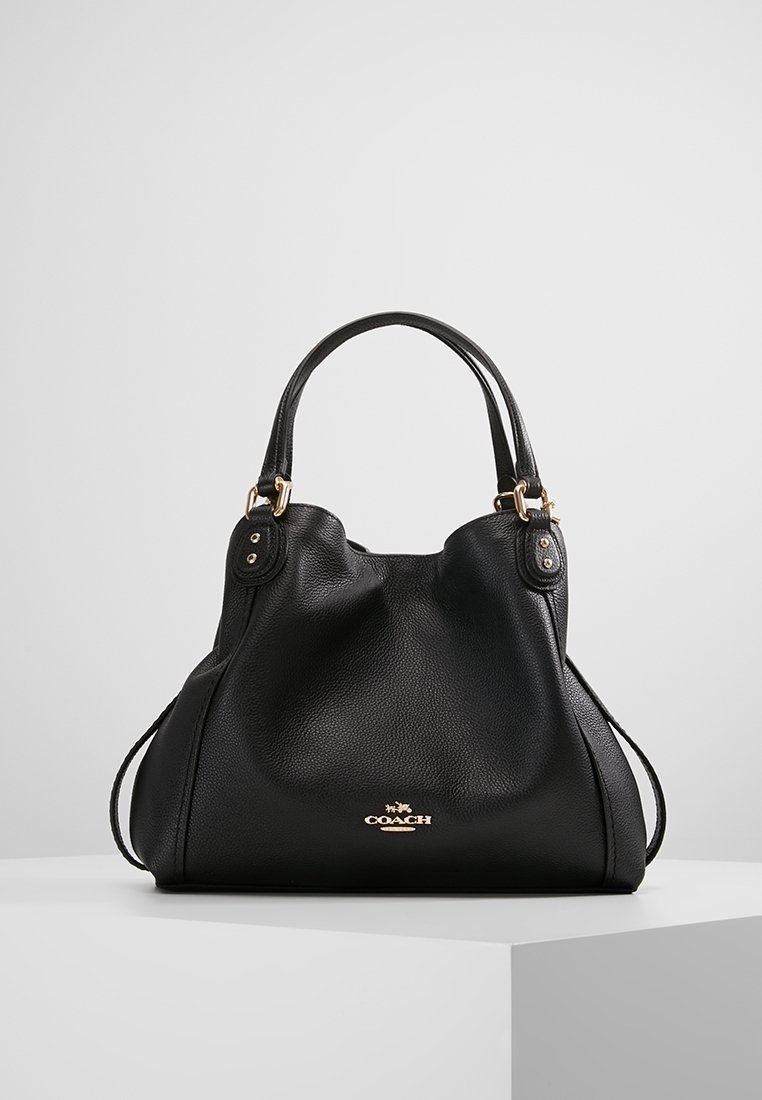 Sac à main Coach en cuir noir avec doubles poignées, ferrures dorées et emblème logo, présenté sur une surface blanche avec un fond gris clair uni.