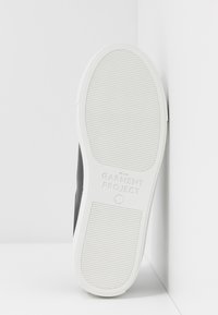 Semelle d'une basket blanche avec des motifs de bande de roulement horizontaux fins, marquage "GARMENT PROJECT" et taille 37, inclinée contre un mur d'angle blanc.