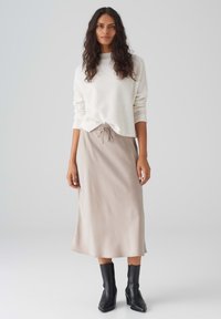 Helle beige Strickpullover mit geripptem Ausschnitt, kombiniert mit einem glatten beigen Midirock mit gerafftem Kordelzugbund und schwarzen Knöchelstiefeln.