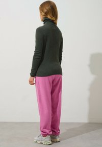 Dunkelgrüner Rollkragenpullover, figurbetont, kombiniert mit lockeren, pastellfarbenen rosa Jogginghosen. Das Model trägt graue Sneaker mit grünen Akzenten.
