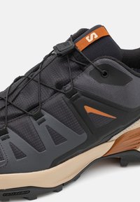 Gros plan d'une chaussure de trail noire et grise avec des touches orange, maintenue par un système de laçage rapide et une semelle beige amortissante.