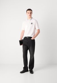 Witte poloshirt met grijze accenten, zwarte rechte jeans, zwarte schoenen. Model houdt een zwarte clutch vast en staat op een neutrale achtergrond.