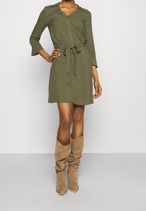 Nainen pukeutuneena oliivinvihreään napilliseen mekkoon, jossa on sidottu vyötärönauha, ja beiget löysät mokkanahkaiset polven korkeudella olevat saappaat, seisoo yksinkertaisen taustan edessä.