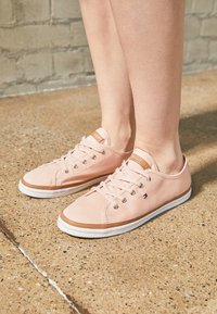 Jambes portant des baskets basses rose pâle avec des semelles et lacets blancs, debout sur un béton texturé couleur fauve près d'un mur en briques.