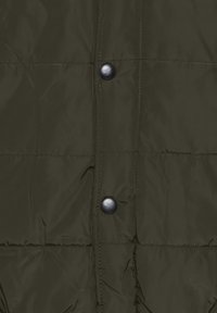 Gros plan d’un tissu matelassé vert foncé avec deux boutons pression noirs alignés verticalement sur la fermeture avant d’une veste ou d’un manteau.