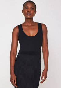 Donna con i capelli corti indossa un abito senza maniche a coste nero, orecchini a cerchio dorati e anelli, in piedi su uno sfondo semplice e chiaro.