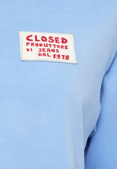 Ανοιχτό μπλε βαμβακερό μπλουζάκι με λευκή ραμμένη ετικέτα που φέρει κόκκινο κεντητό κείμενο: «CLOSED PRODUTTORE DI JEANS DAL 1978.»
