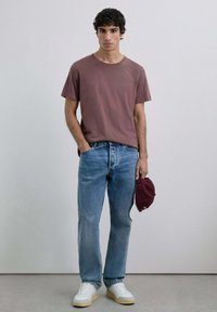 Maronfarvet bomulds-T-shirt, lyseblå denimjeans, hvide sneakers med beige såler, holder en maronfarvet kasket. Simpelt design, afslappet pasform.