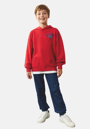 Smilende dreng iført en rød hættetrøje med teksten "Great Day", blå bukser og hvide sneakers mod en enkel lys baggrund.