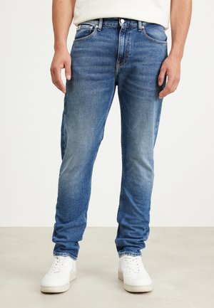 Vaqueros de mezclilla azul claro con corte recto, cinco bolsillos y sutiles detalles de desgaste, combinados con zapatillas blancas.