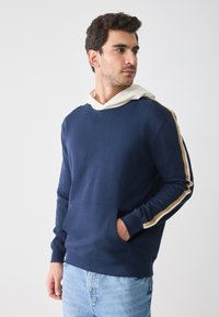 Felpa navy con cappuccio, dotata di tasca frontale e cordino bianco, con strisce dorate e bianche sulle maniche e vestibilità comoda.