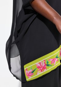 Top negro fluido con mangas cortas, que presenta un estampado floral verde y rosa en el dobladillo y un panel lateral transparente para añadir textura.