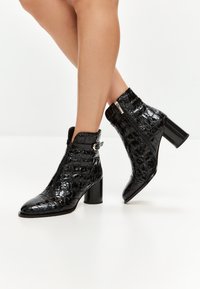 Schwarze, glänzende Knöchelstiefel mit Krokoprägung, Blockabsätzen, seitlichen Reißverschlüssen und verstellbaren Riemen, getragen an übergeschlagenen Beinen vor weißem Hintergrund.
