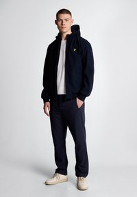 Μπλε σκούρο φούτερ με φερμουάρ και κουκούλα, με μικρό κίτρινο λογότυπο, σε συνδυασμό με μπλε σκούρο παντελόνι και μπεζ αθλητικά παπούτσια, πάνω σε μοντέλο μπροστά σε απλό φόντο.