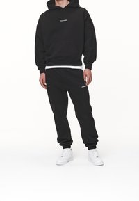 Sudadera negra con capucha, bolsillo frontal y texto blanco; combinada con joggers negros a juego que presentan una marca blanca, usados con zapatillas blancas.