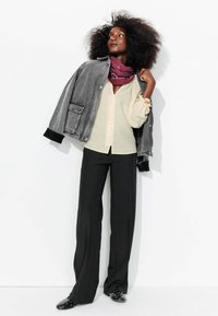 Veste en denim gris avec des poignets noirs, chemise beige à boutons, pantalon large noir et chaussures noires à enfiler avec des clous, accessoirisé avec une écharpe bordeaux.