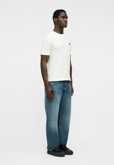 Μαύρος άντρας που στέκεται φορώντας μπλε τζιν, μαύρα αθλητικά παπούτσια και λευκό κοντομάνικο μπλουζάκι με ένα μικρό κεντημένο κεφάλι τίγρης στο στήθος.