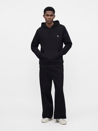 Homme debout les mains dans les poches, portant un sweat à capuche noir, un pantalon noir ample et des baskets blanches sur un fond blanc uni.