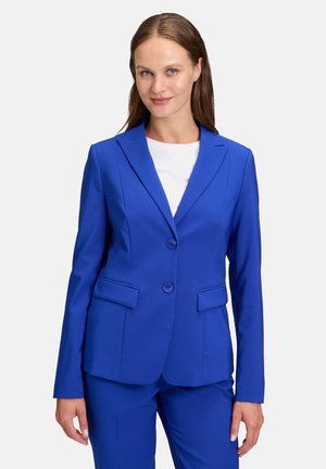 Blazer blu con rever a punta, chiusura a due bottoni e tasche laterali, realizzato in un tessuto liscio; abbinato a una maglietta bianca sotto.