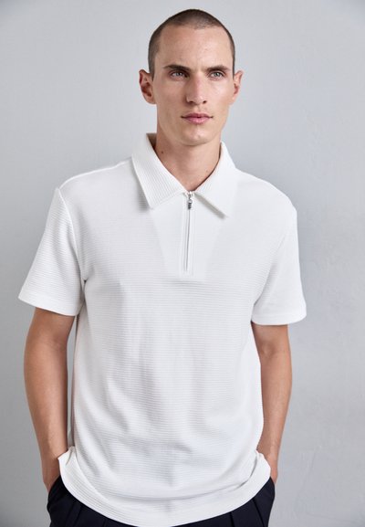 Jeune homme aux cheveux courts portant un polo à manches courtes blanc avec une fermeture éclair à l'avant, se tenant devant un mur gris clair uni.