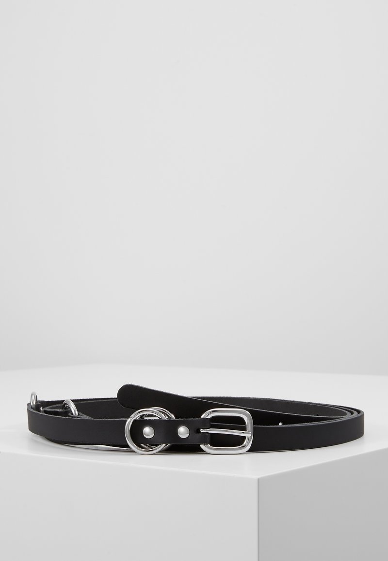 Ceinture en cuir noire avec boucle argentée et anneaux métalliques ronds, présentée enroulée sur une surface blanche sur fond uni.