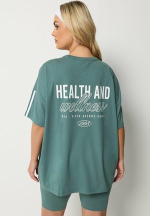 Kobieta z długimi blond włosami, ubrana w zielony oversize'owy t-shirt i zestaw szortów z napisem „Zdrowie i dobre samopoczucie” na plecach, biała opaska na głowę.