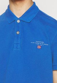 Polo bleu avec deux boutons argentés, texte blanc brodé "NAPAPIJRI" et petit drapeau norvégien sur la poitrine.