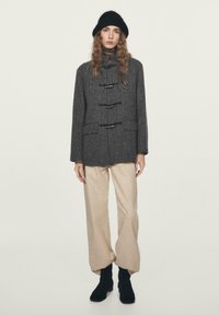 Cappotto grigio in lana con chiusure a fibbia, abbinato a pantaloni beige a gamba larga e stivaletti neri alla caviglia. La modella indossa un cappello di lana nero.