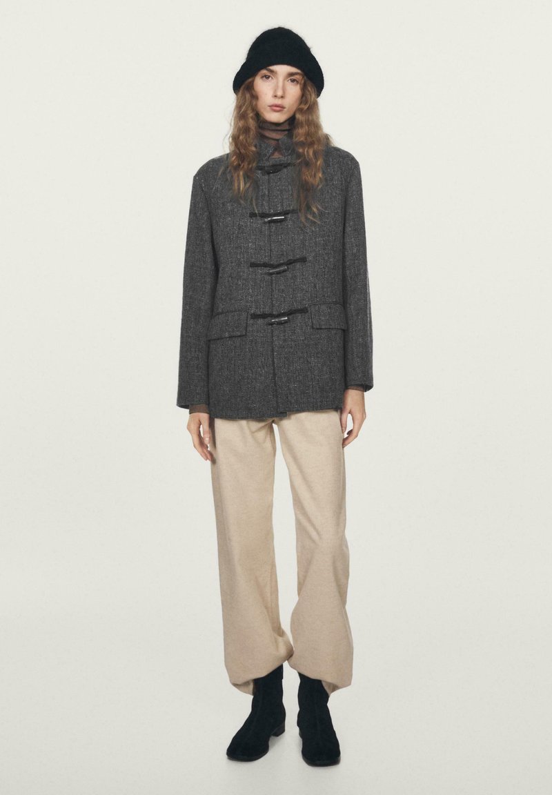 Cappotto grigio in lana con chiusure a fibbia, abbinato a pantaloni beige a gamba larga e stivaletti neri alla caviglia. La modella indossa un cappello di lana nero.