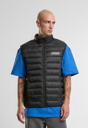 Gilet nero imbottito con colletto alto e zip frontale. Presenta un patch con logo ed è indossato sopra una maglietta a maniche corte blu, abbinato a pantaloni grigi.