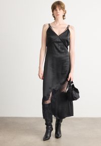 Schwarzes Satin-Kleid mit einer Spitzenbordüre, dünnen Trägern und einer glatten Textur. Kombiniert mit einer schwarzen Stoffhandtasche und Ankle Boots.
