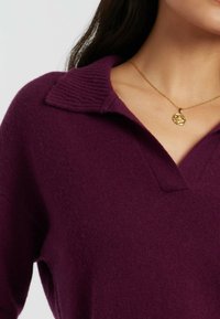 Strickpullover in tiefem Lila mit einem gerippten Kragen und einem V-Ausschnitt-Design. Trägt eine goldene Anhänger-Kette. Glatte Textur mit einem lockeren Schnitt.