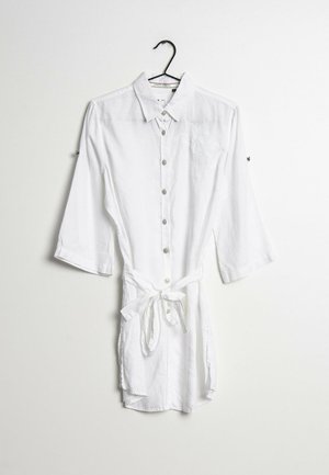 Robe chemise blanche à manches longues, boutonnée, avec col et ceinture nouée à la taille, suspendue sur un cintre noir sur fond blanc.