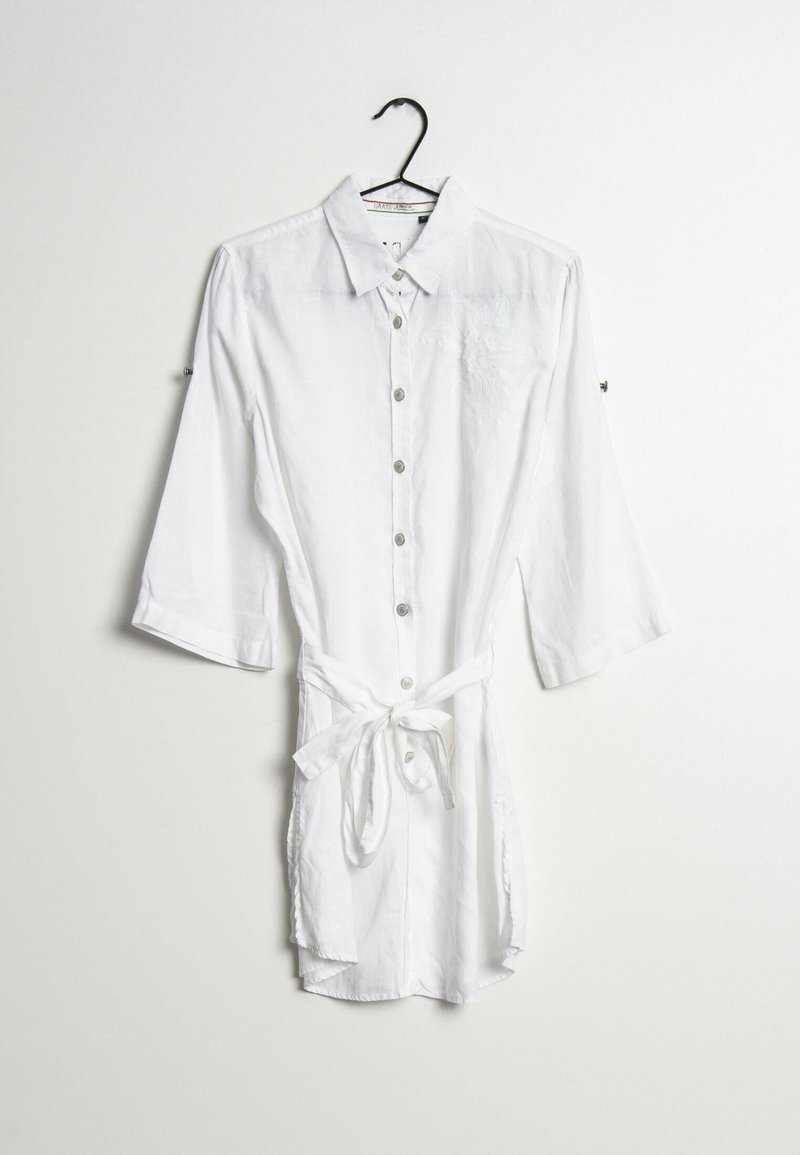Robe chemise blanche à manches longues, boutonnée, avec col et ceinture nouée à la taille, suspendue sur un cintre noir sur fond blanc.